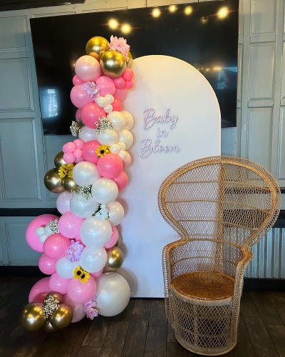 A pink, white and gold balloon garland around a “Baby in Bloom” arch, with a wicker chair